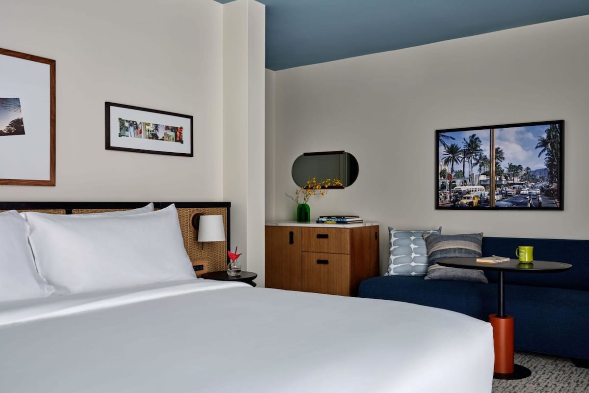 A hotel room with a bed, a sofa, a small table, a mirror, and two framed pictures on the wall.