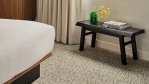 Habitación de hotel con cama, armario de madera, ventana que ofrece vistas a edificios y palmeras, y una mesita con un jarrón de flores y libros.