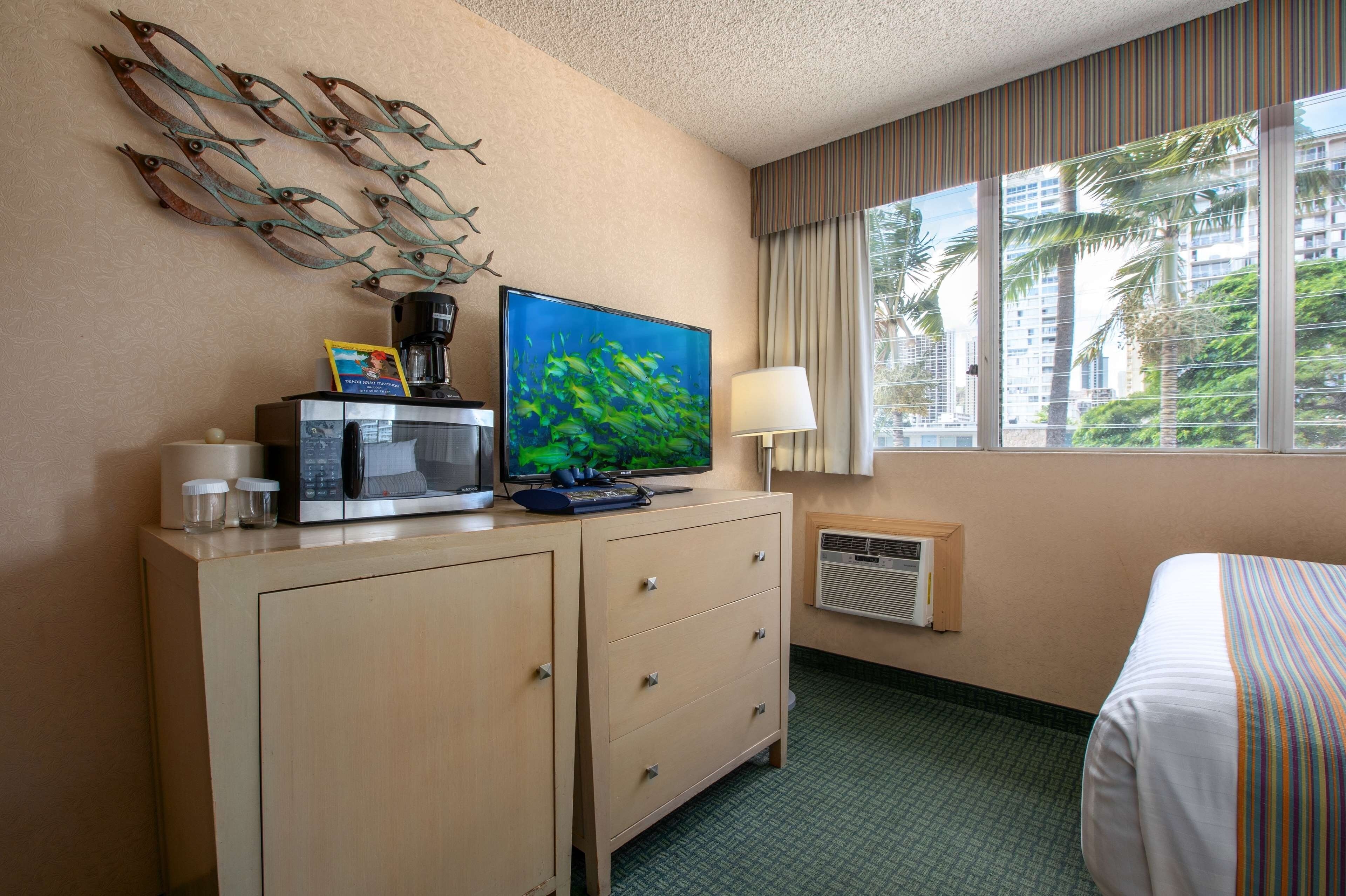 A hotel room with a bed, a desk with a television, a coffee maker, and a window with a view of palm trees and buildings.