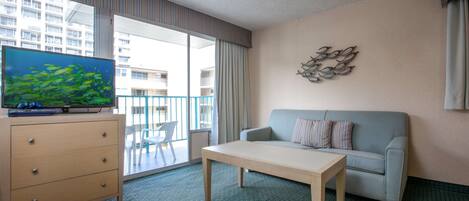 A hotel room with a sofa, a wooden table, a TV on a dresser, and a balcony with a view of buildings.