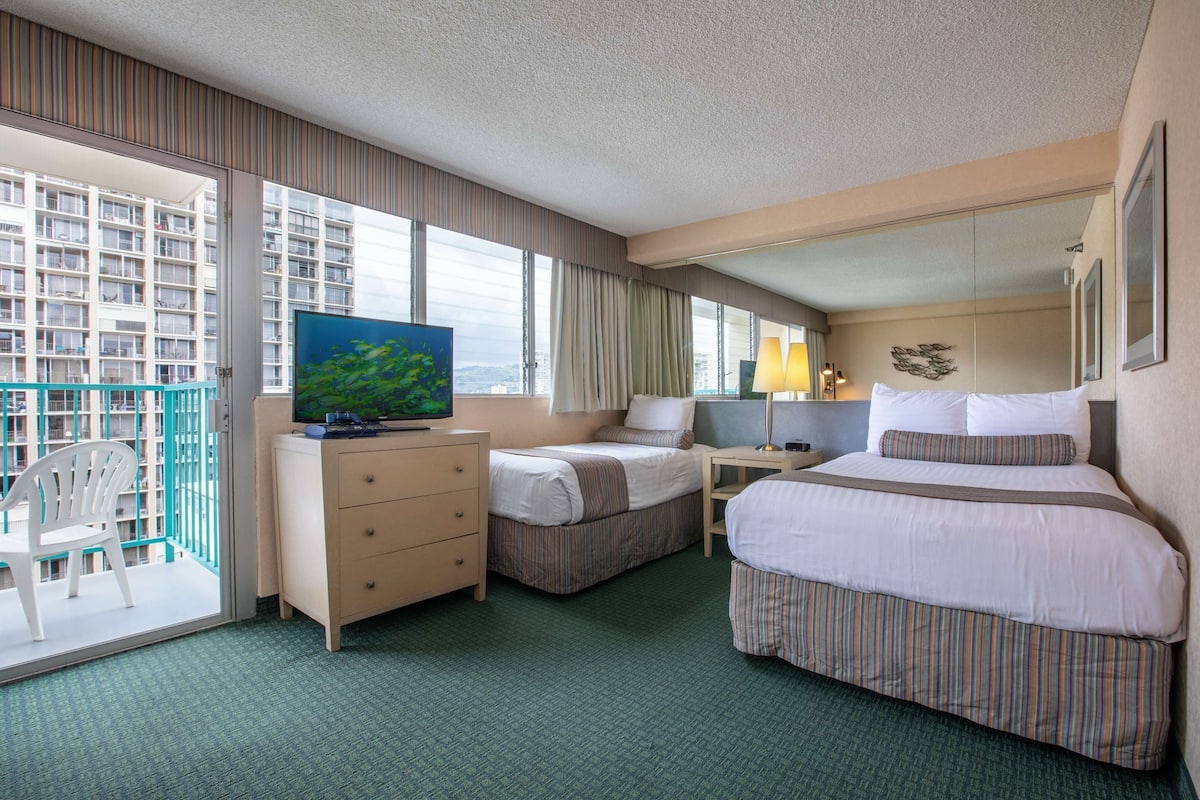Een hotelkamer met twee bedden, een televisie, een bureau en een balkon met uitzicht op de stad.