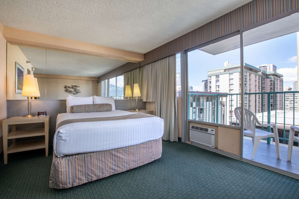 Een hotelkamer met een groot bed, twee bedlampen, een klein tafeltje, een stoel en een balkon met uitzicht op gebouwen.