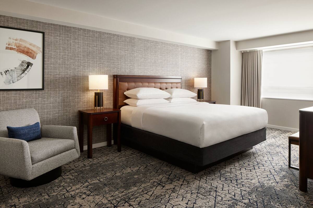 A hotel room with a large bed, a chair, two bedside tables with lamps, a framed artwork on the wall, and a window with curtains.