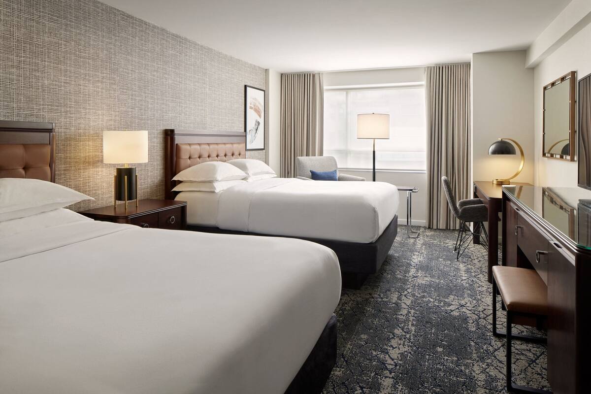 A hotel room with two beds, a desk, a chair, a lamp, and a large window with curtains.