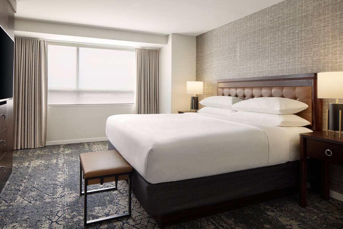 A hotel room with a large bed, a bench, two bedside tables with lamps, a TV, and a large window with curtains.