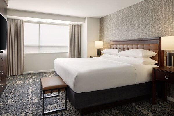 A hotel room with a large bed, a bench, two bedside tables with lamps, a TV, and a large window with curtains.