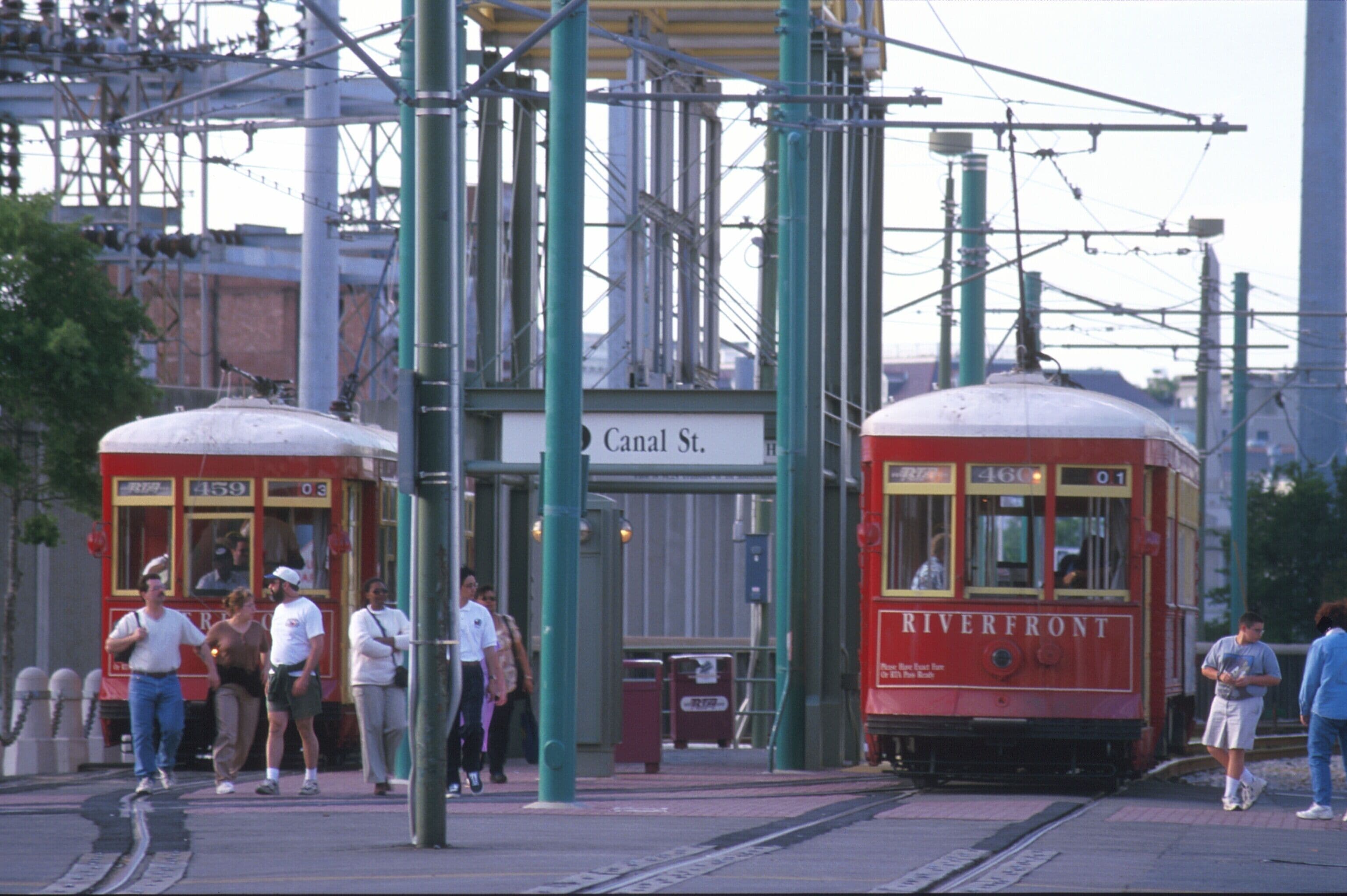 Une station de tramway où deux tramways sont arrêtés, portant les indications « Riverfront » et « Canal St. ». Des personnes attendent près du quai.