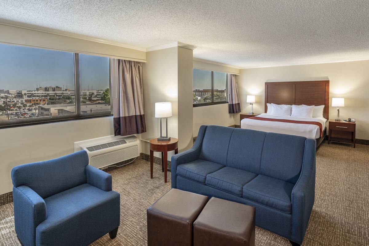 A hotel room with a large bed, a sofa, a chair, a TV, and a view of the city through large windows.