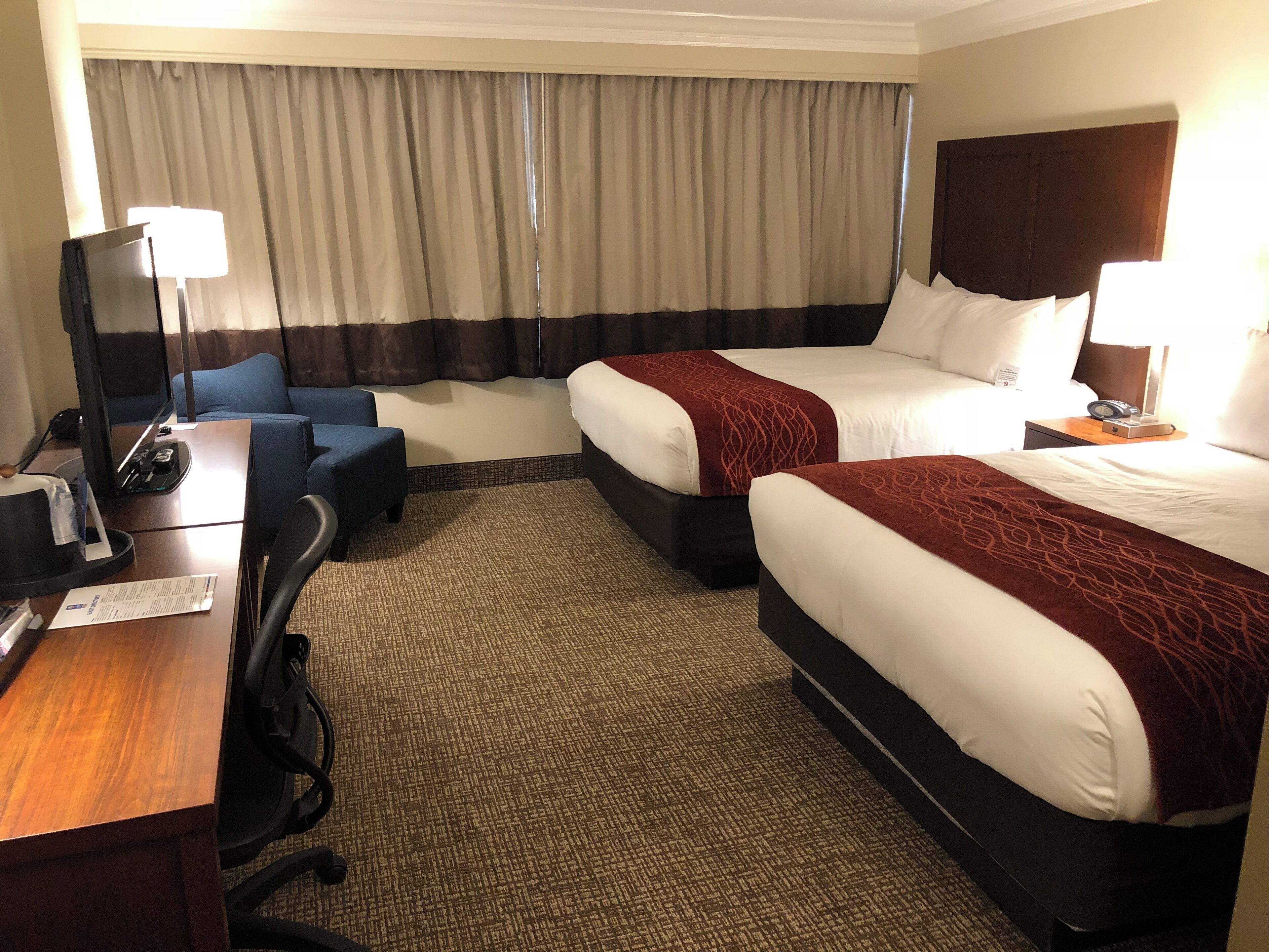 A hotel room with two beds, a desk, a chair, a lamp, and a TV.