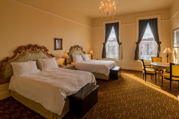 A hotel room with two beds, a chandelier, a dining table with chairs, and large windows with curtains.