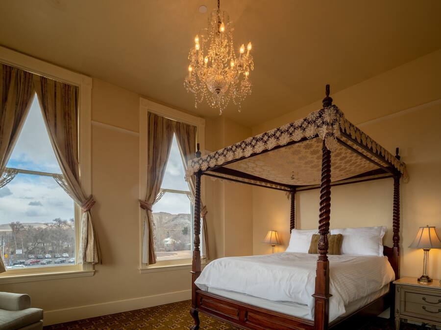 Una cama con dosel adornada con un candelabro, vistas de la ciudad a través de amplias ventanas y una mesita de noche con lámpara.