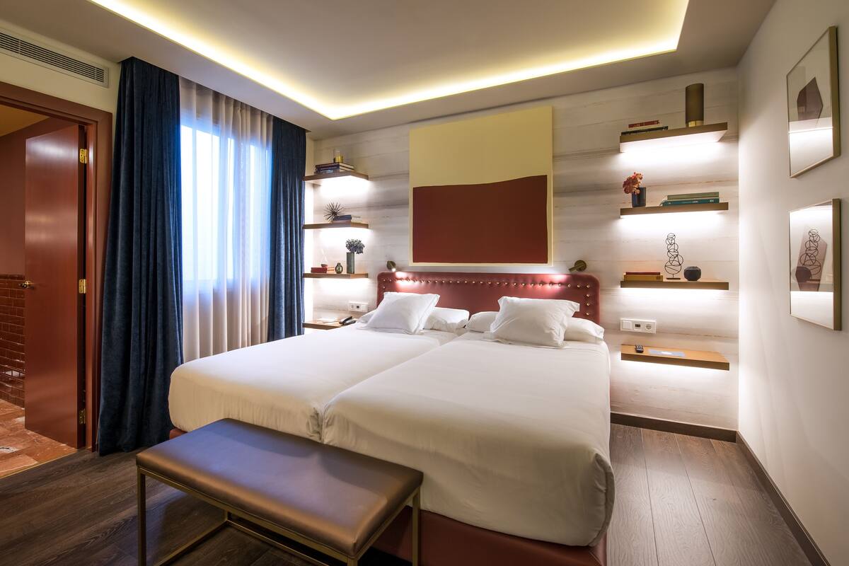 A hotel room with a double bed, bedside tables, a bench, and a large window with curtains.