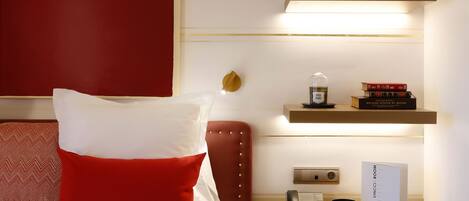 A hotel room with a bed, a red pillow, a nightstand with a phone, a vase with flowers, and books on shelves.