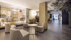 Un hall d’entrée moderne avec un comptoir de réception en bois, un espace salon avec des tables rondes et un mur décoratif agrémenté d’étagères.