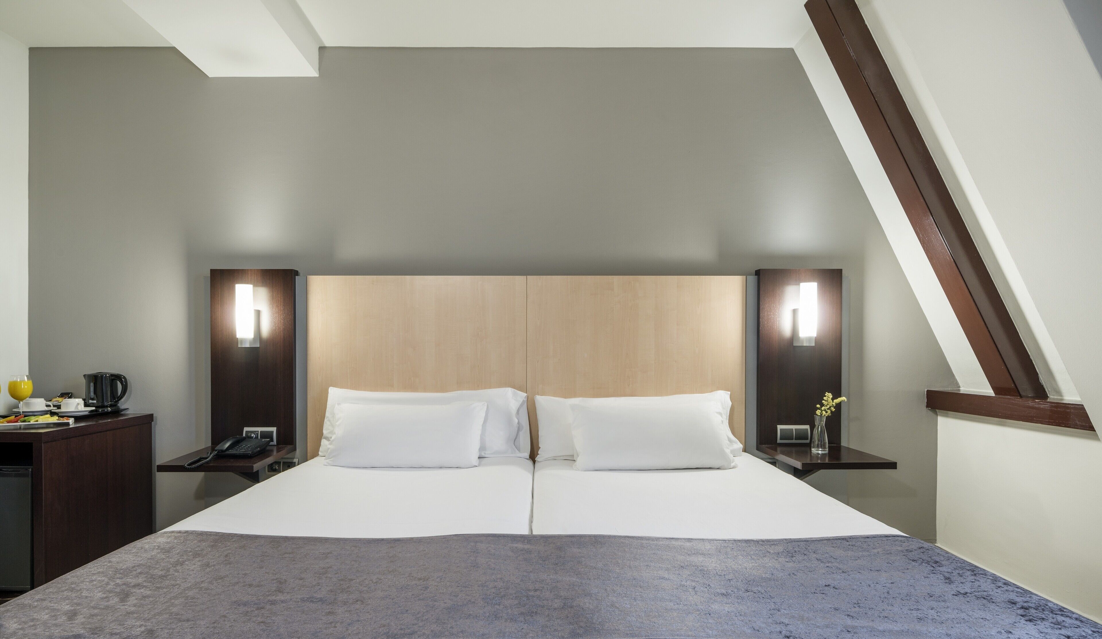 A modern hotel room with a large bed, two bedside tables, a nightstand, a desk with a lamp, and a small table with a vase.