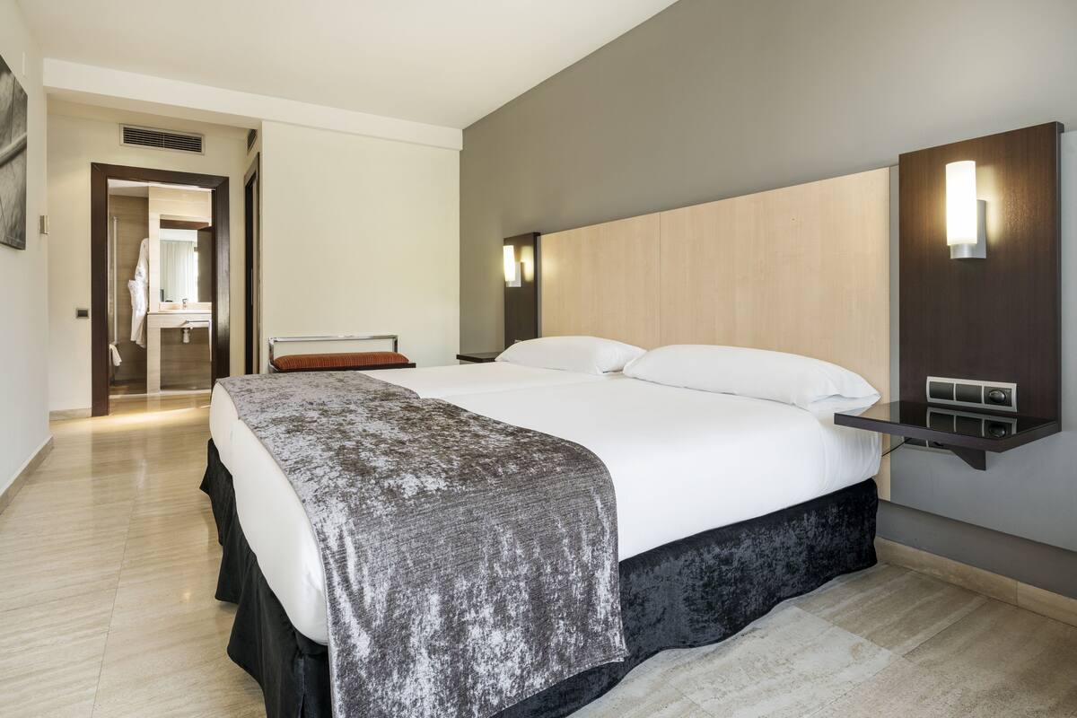 A modern hotel room with a large bed, a wooden headboard, a bedside table, and a wall-mounted light.