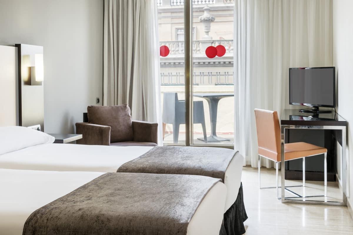 A hotel room with two beds, a desk with a TV, a chair, and a view of a balcony with red decorations.