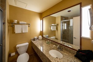 A hotel bathroom with a granite countertop, a white sink, a large mirror, and a shower area.