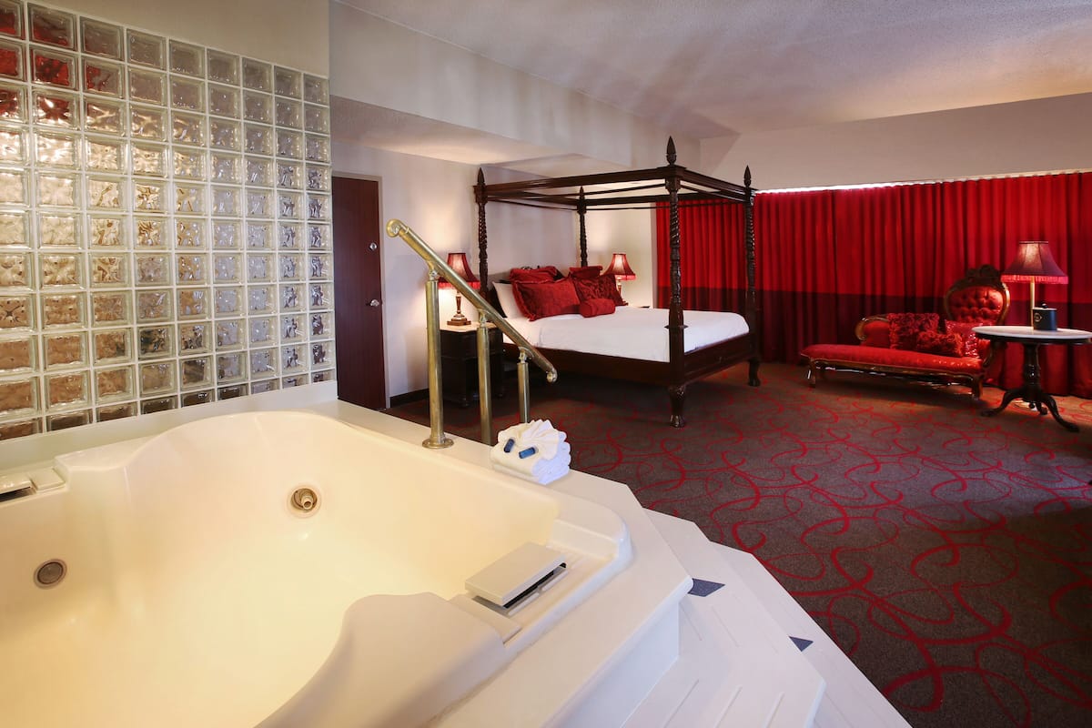 A hotel room with a large white bathtub, a four-poster bed with red bedding, a red carpeted floor, and a red curtain.