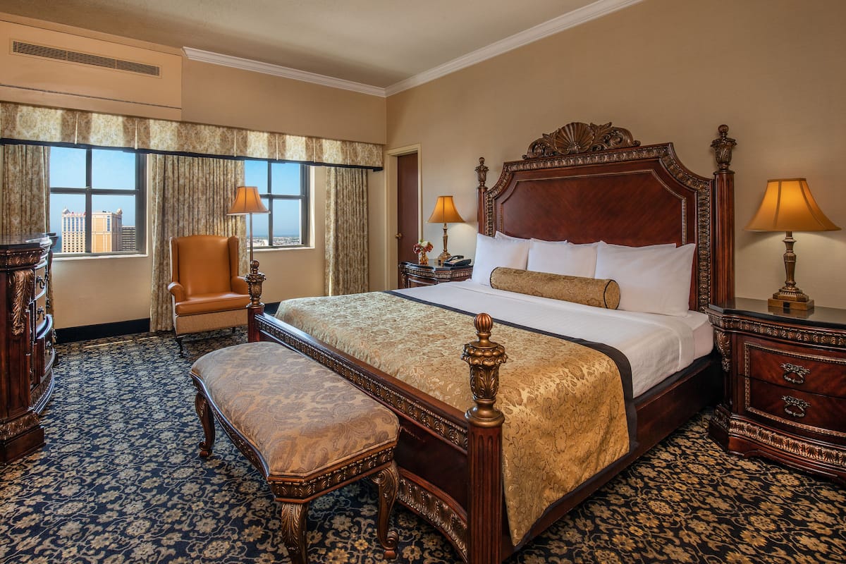 A large bed with a wooden headboard, a bench, a chair, two bedside tables, and a large window with curtains.
