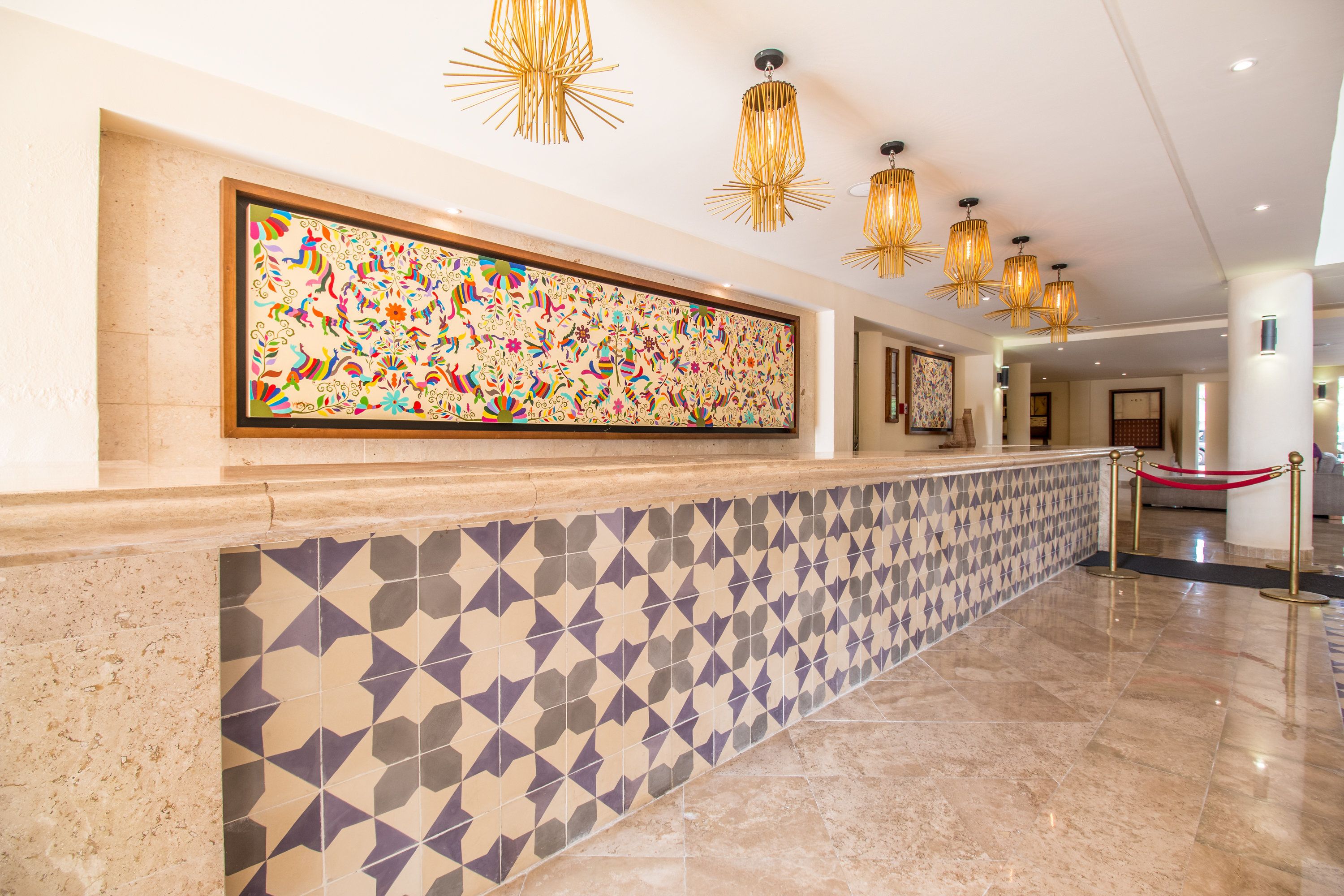 Un hall d’accueil d’hôtel avec un mur recouvert de carreaux, une grande œuvre d’art colorée et de nombreuses lampes suspendues.