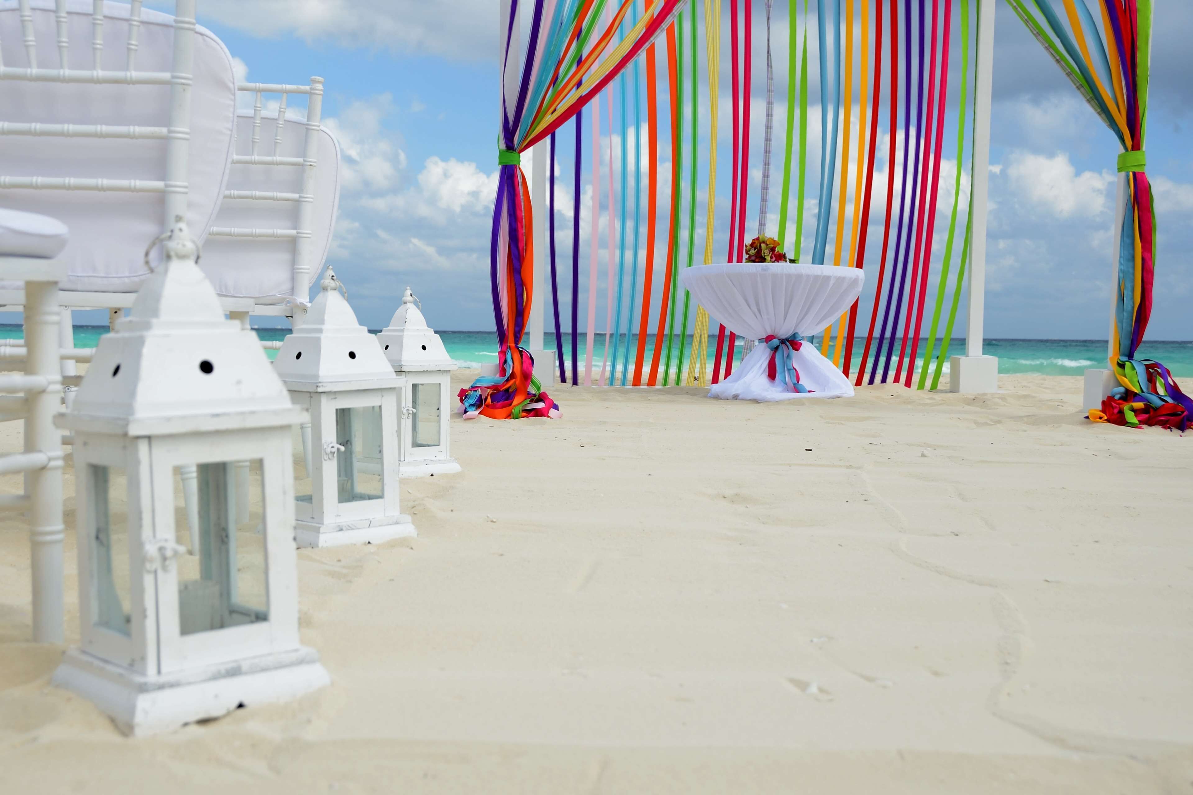 A beach setting with white lanterns, colorful drapes, and a white table with a flower arrangement.
