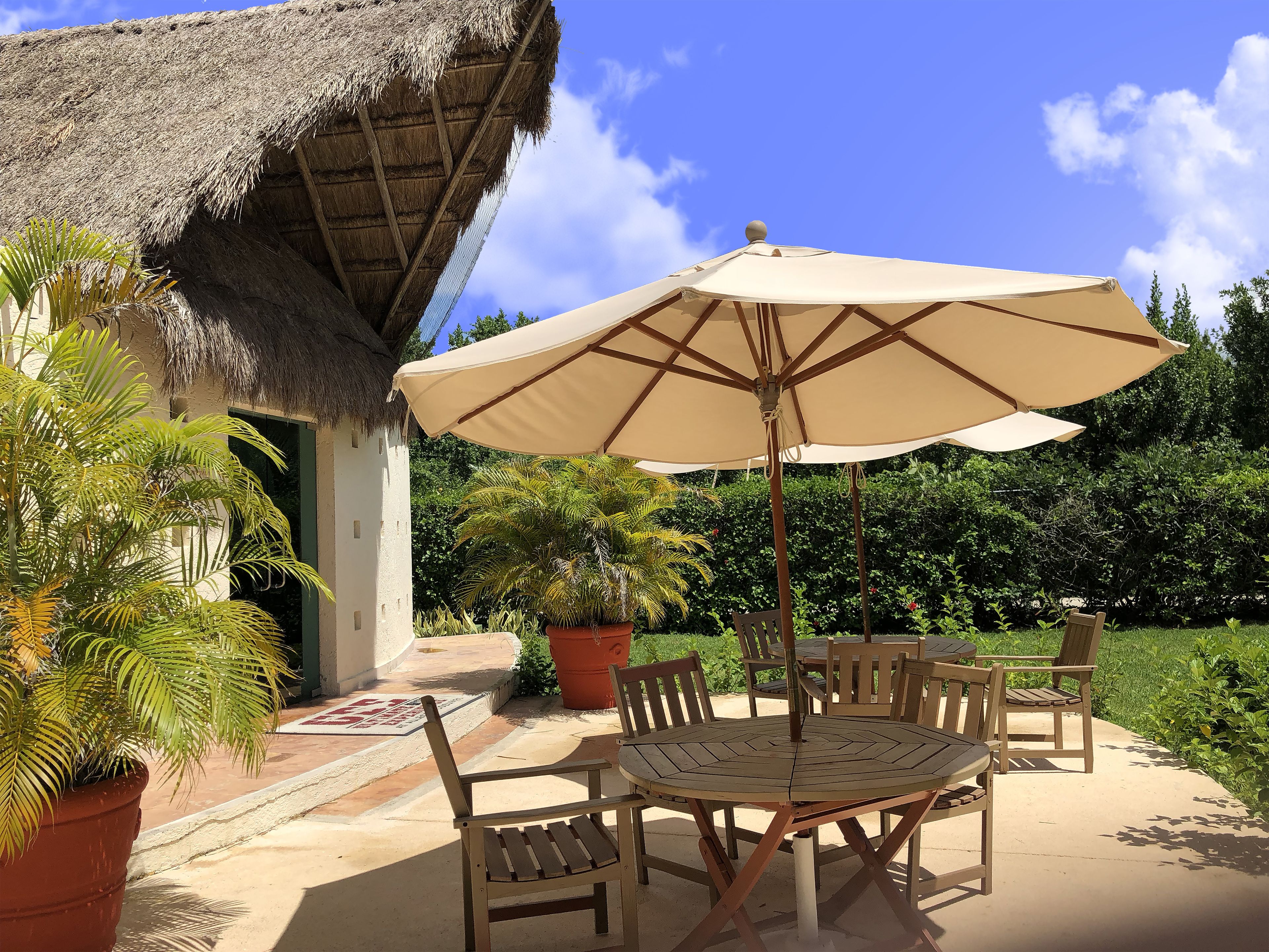 Un patio al aire libre con una mesa y sillas de madera redonda, un paraguas color beige y una estructura con techo de paja.