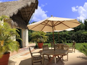An outdoor patio with a round wooden table and chairs, a beige umbrella, and a thatched-roof structure.