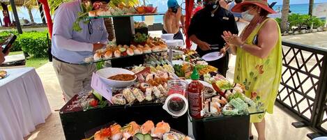 Un bufé con diversos platos, entre ellos sushi y ensaladas, bajo un toldo rojo con palmeras al fondo.