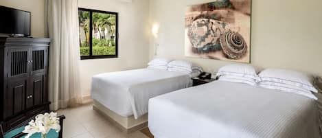 A hotel room with two beds, a TV, a desk with a vase of flowers, a ceiling fan, and a window with a view of greenery.