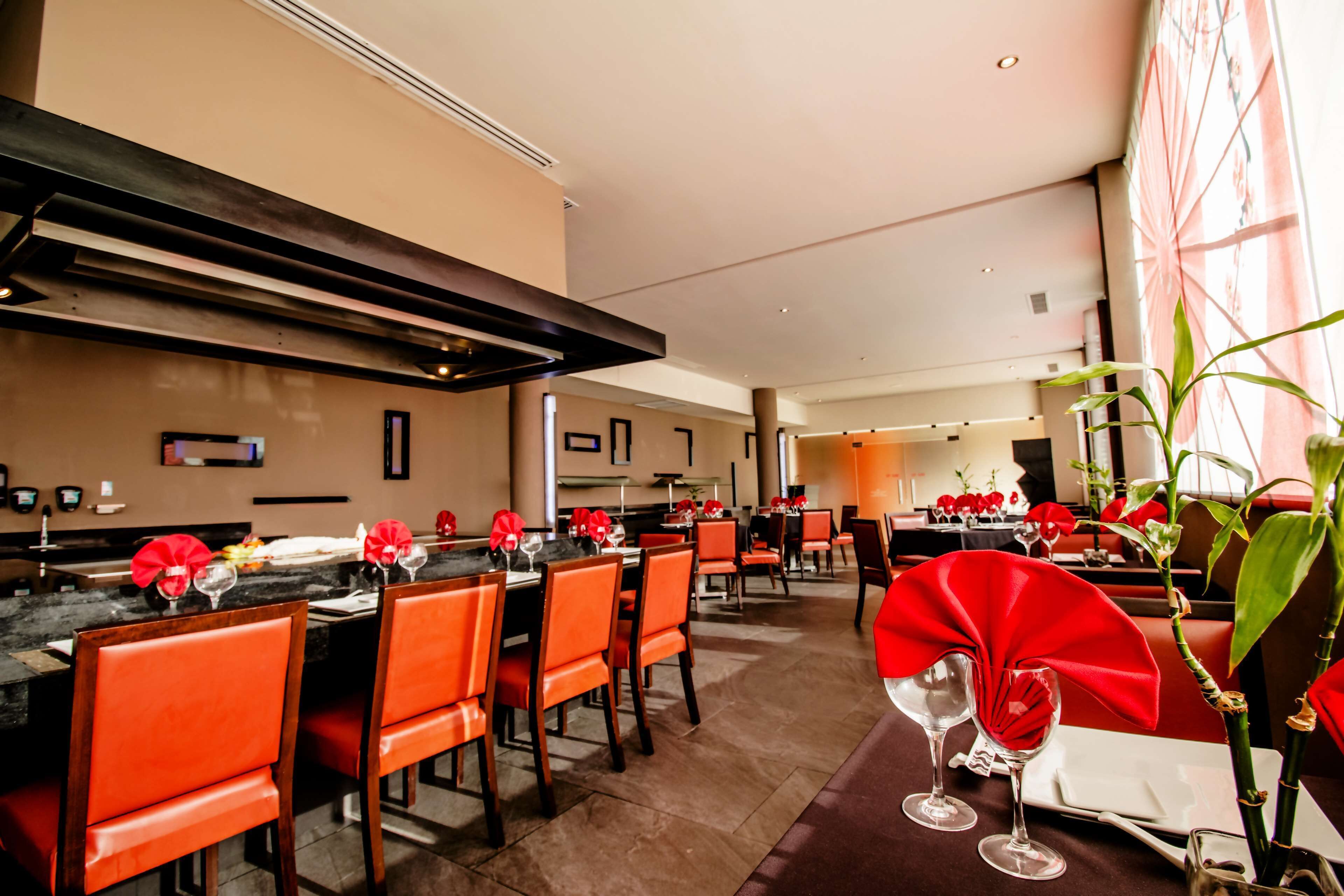 A restaurant with a modern interior, featuring orange chairs, tables set with red napkins, and a large plant in the foreground.