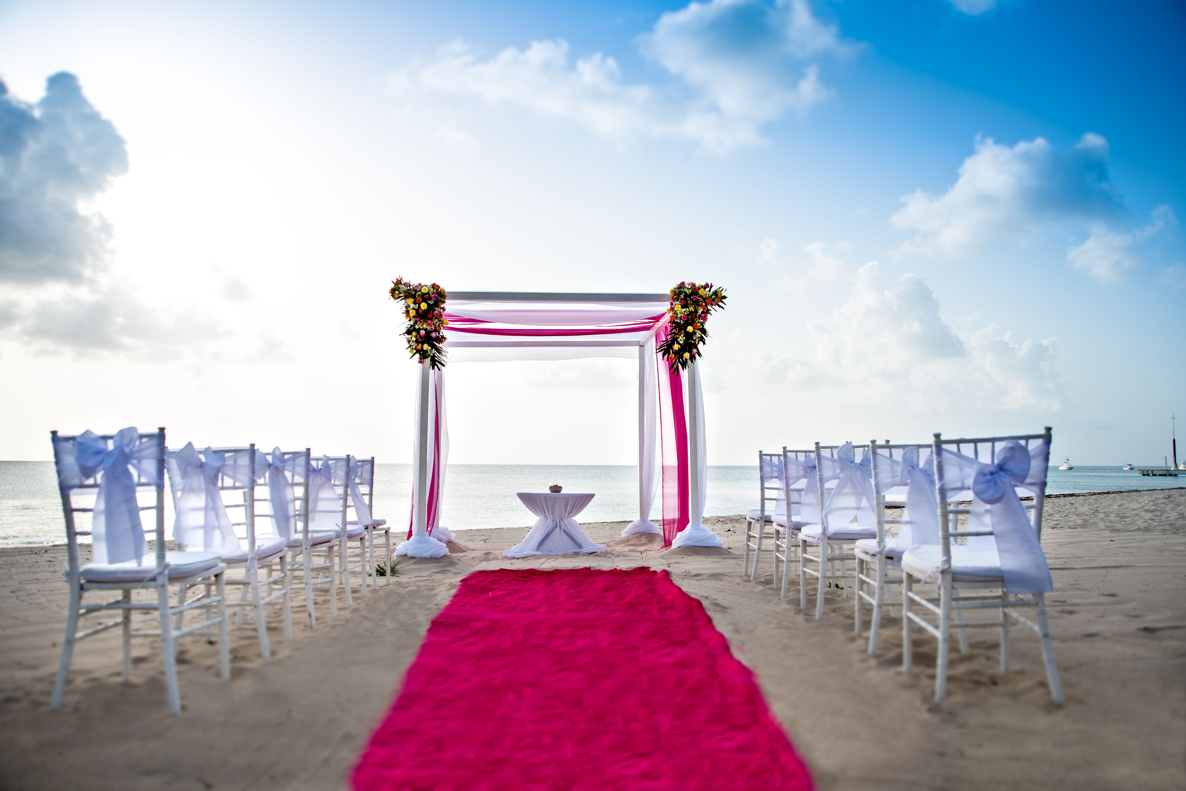 Decoração de casamento na praia, com cadeiras brancas e um tapete vermelho que leva a um arco branco enfeitado com flores.