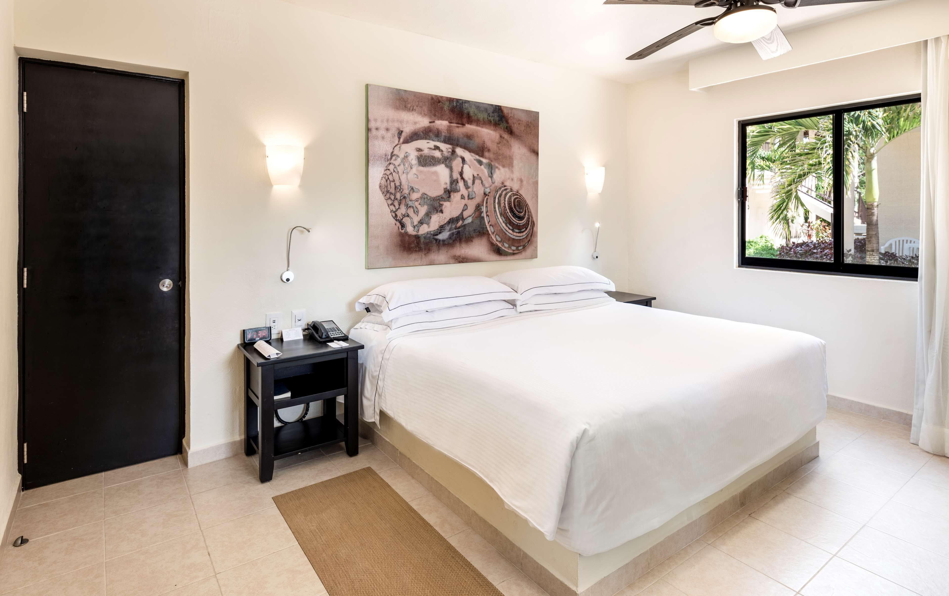 Habitación de hotel con una cama, una mesita de noche con un teléfono y una lámpara, un cuadro en la pared y una ventana con vistas a la vegetación.
