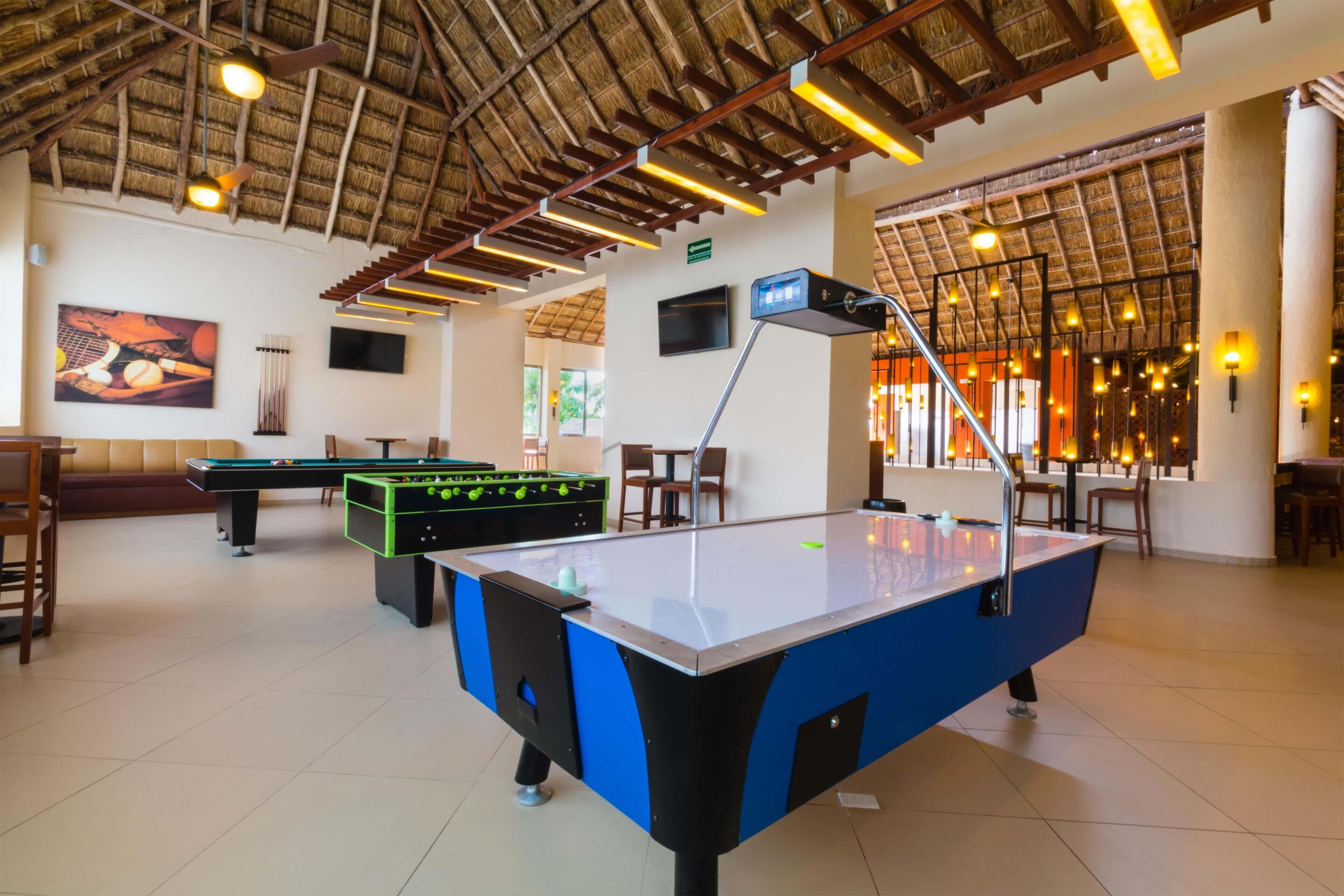 A pool table, foosball table, and billiards table in a well-lit indoor space with a thatched roof ceiling.