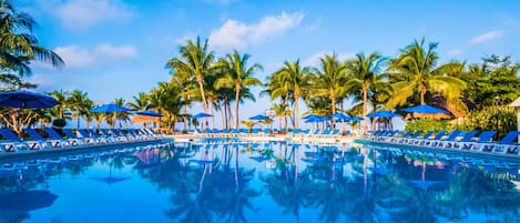 Une piscine bordée de chaises longues bleues et d’ombrelles, le tout entouré de palmiers.