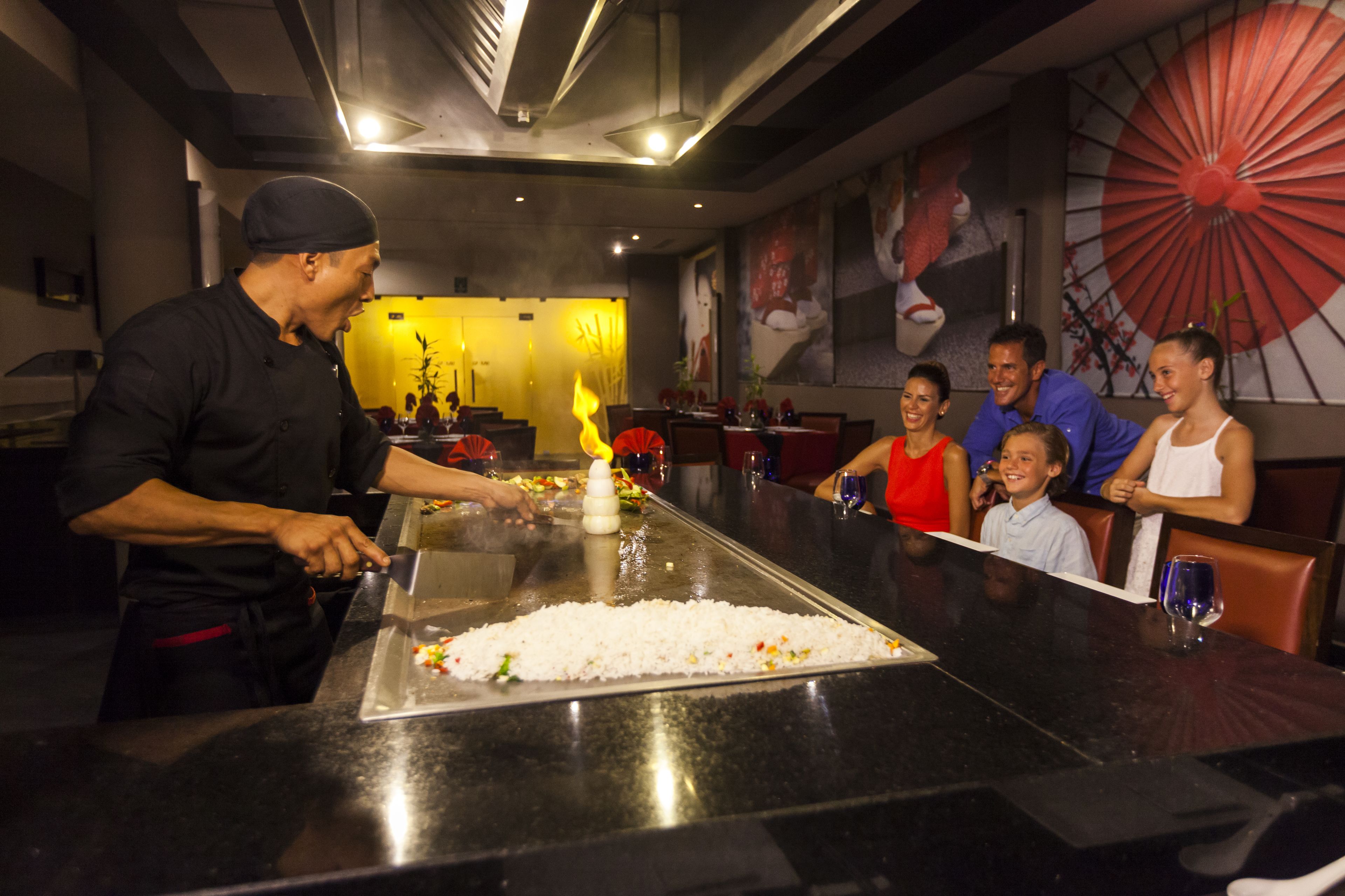 A restaurant with a long counter, a chef preparing food, and diners seated at tables.