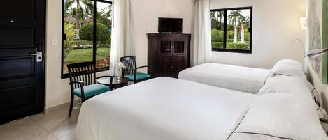 A hotel room with two beds, a ceiling fan, a TV on a cabinet, and a view of the garden through the window.