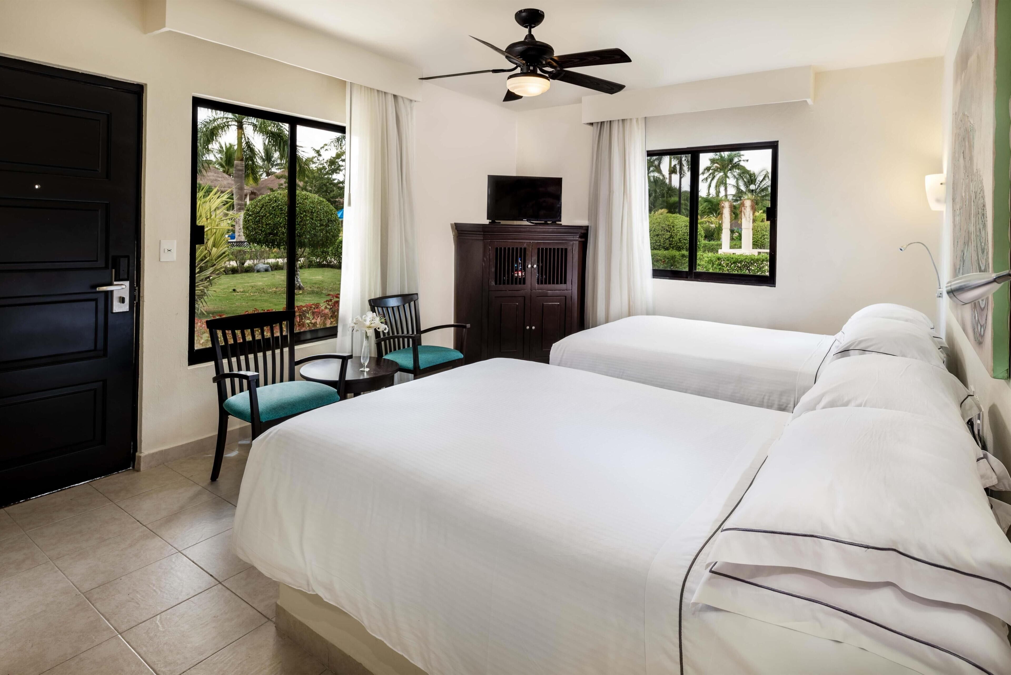Une chambre d’hôtel avec deux lits, un ventilateur de plafond, un téléviseur posé sur un meuble et une vue sur le jardin grâce à de grandes fenêtres.