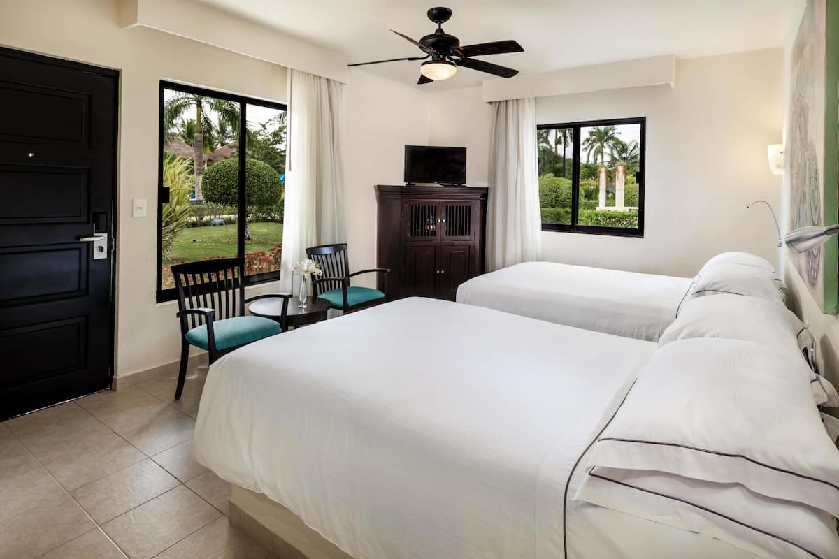 Habitación de hotel con dos camas, un ventilador de techo, un televisor en un mueble y vistas al jardín a través de ventanas amplias.