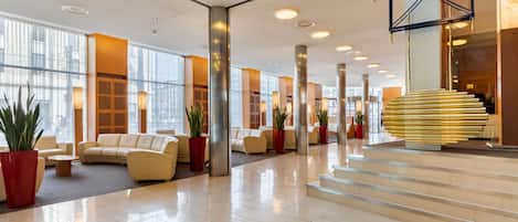 Un lobby espacioso con diseño moderno, que cuenta con una escalera central, zonas de estar cómodas y amplios ventanales que permiten la entrada de luz natural.