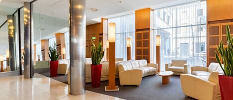 A modern lobby with a spacious layout, featuring a central seating area with sofas and a coffee table, surrounded by large windows and potted plants.