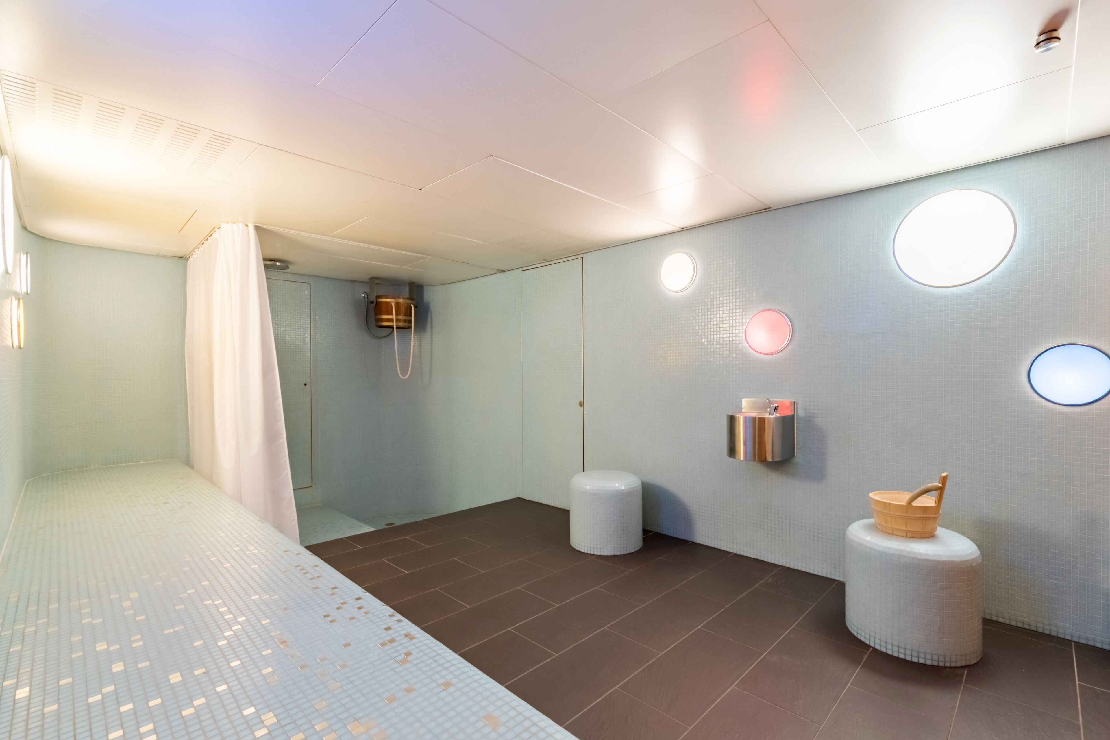 Ein modernes Badezimmer mit Dusche, Toilette und Waschbecken. An der Wand befinden sich zwei runde Spiegel und ein kleiner Hocker mit Korb.