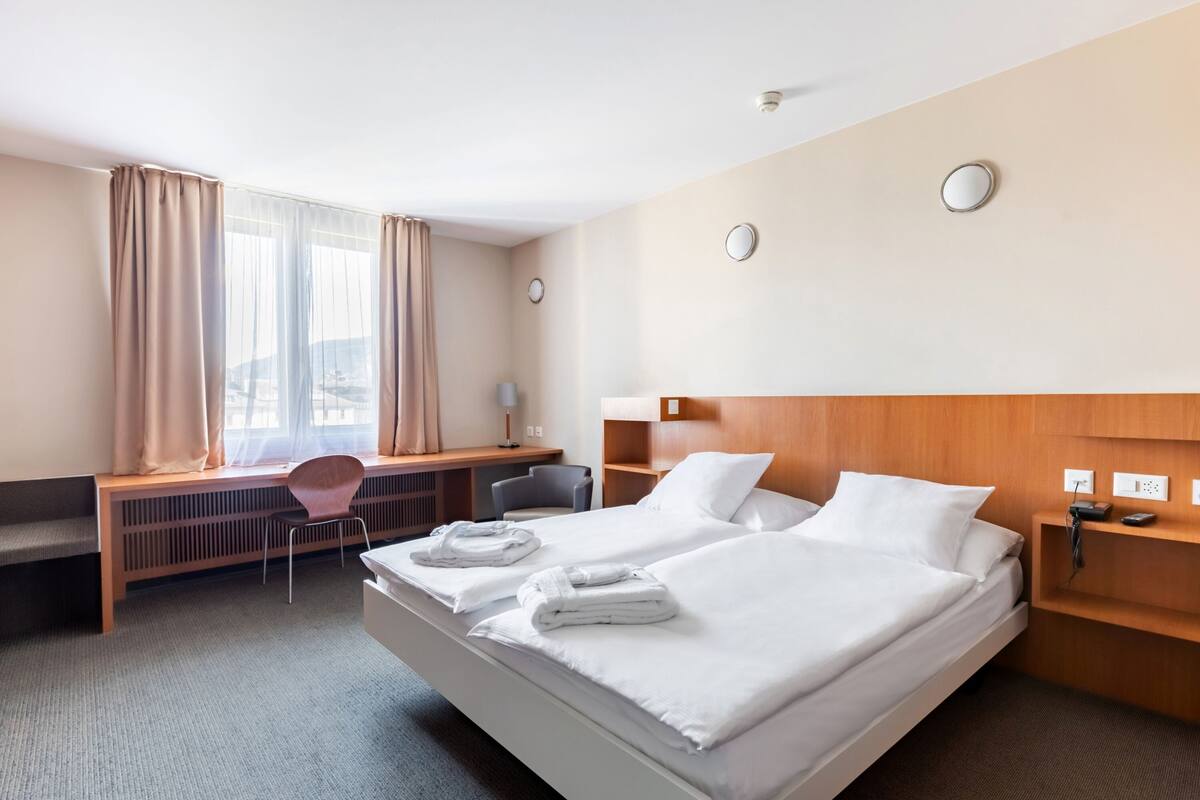 Habitación de hotel con dos camas, un escritorio, una silla, un radiador y una ventana con cortinas.