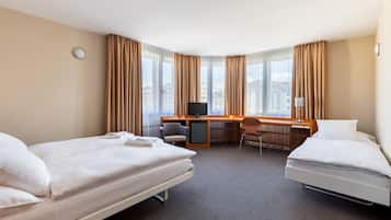 Habitación de hotel con dos camas, un escritorio, una silla, un televisor y ventanas grandes con cortinas.