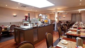 A hotel dining area with multiple tables set for breakfast, a central buffet station, and a large window providing natural light. - Drake Longchamp (Geneva)