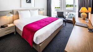A hotel room with a bed, bedside table, lamp, chair, desk, and a window with curtains. - Drake Longchamp (Geneva)