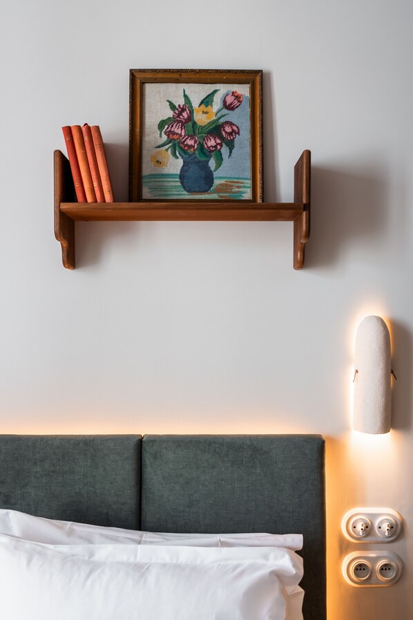A neatly arranged bed with a headboard, a bedside table with a lamp, and a wall-mounted shelf with books and a framed artwork. - Kraft Hotel (Paris)