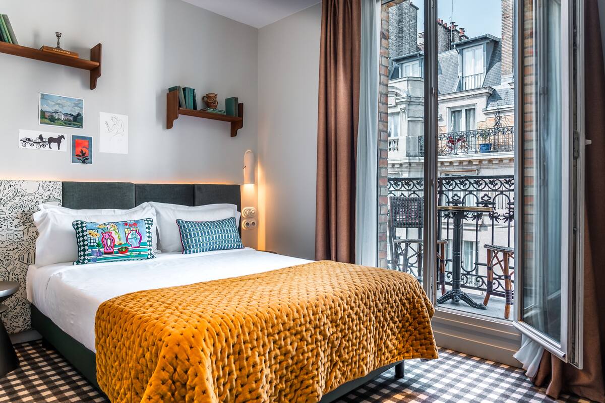 A hotel room with a large bed, a view of the city through a window, and a balcony with outdoor furniture.