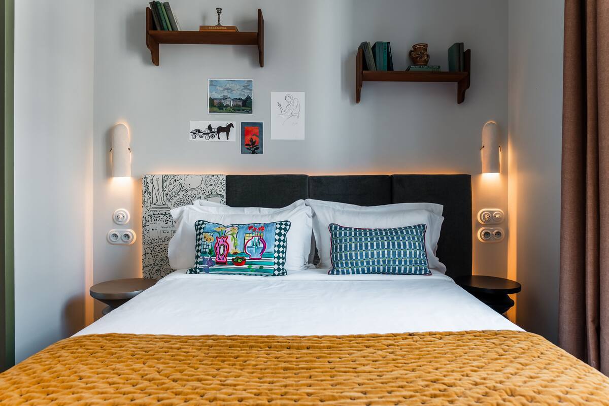 A neatly made bed with a patterned blanket, two bedside tables with lamps, and wall-mounted shelves with books and decorative items.