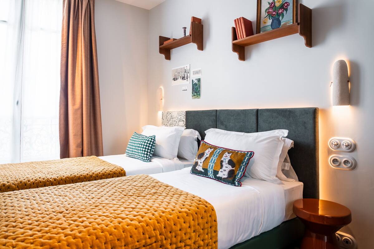 A hotel room with two beds, a large window with curtains, wall-mounted shelves with books and pictures, and a bedside table with a lamp.